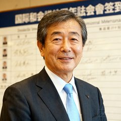 一位五十多岁的日本男性医生半身照，穿着深色西装搭配浅蓝色领带，背景为国际会议签到板，神情专注且友善。